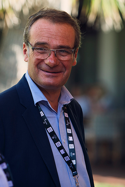 9431-2 Retrato de un hombre con gafas, traje y acreditación colgada al cuello, sonriendo en un entorno exterior con luz natural.